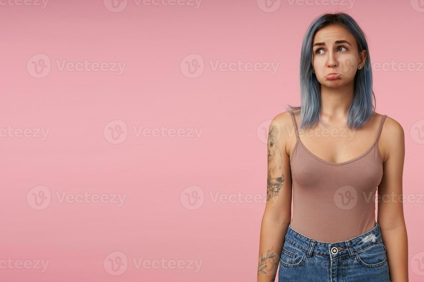 Studio shot of young upset short haired tattooed lady twisting sadly her mouth while looking aside and keeping hands along body, standing over pink background photo