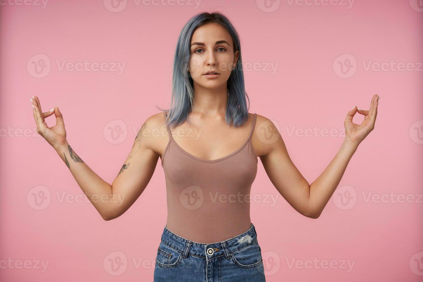 estudio Disparo de atractivo joven hembra con corto azul pelo plegable con elevado dedos namaste firmar mientras en pie terminado rosado antecedentes en beige camisa foto