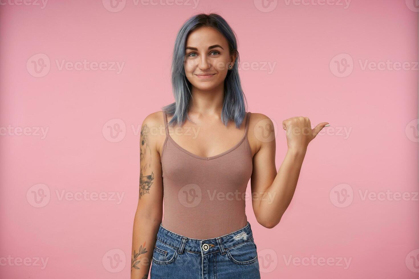 Positive young lovely tattooed lady with short blue hair smiling pleasantly while showing aside with raised hand, being isolated over pink background in casual wear photo