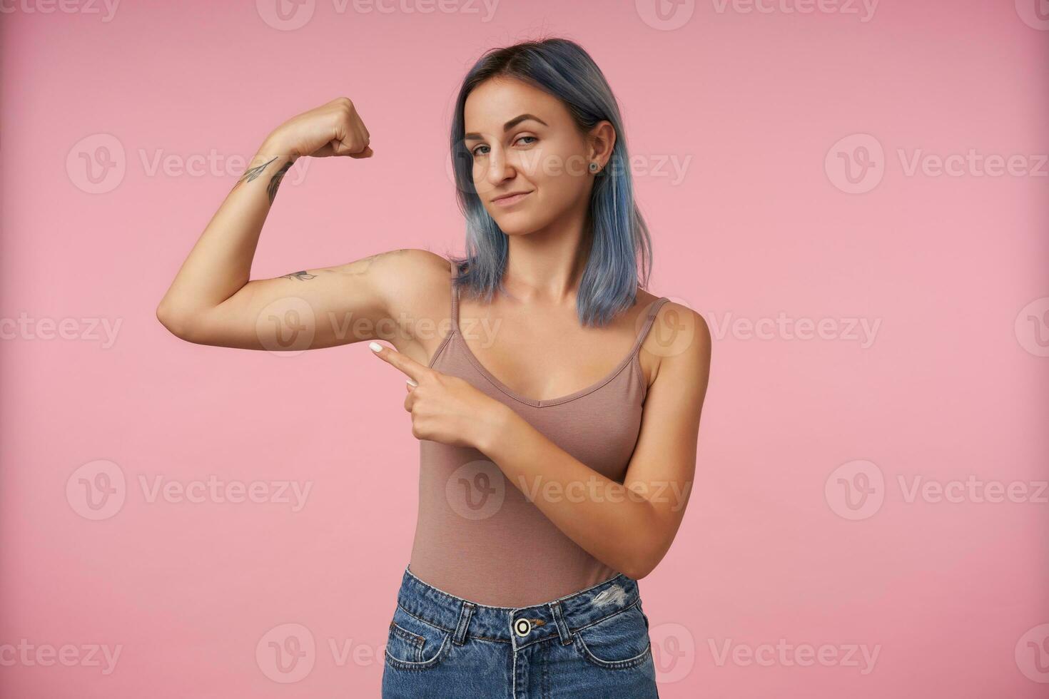 Portrait of young attractive tattooed female with short haircut pointing on her raised hand while showing her strong biceps, standing over pink background photo