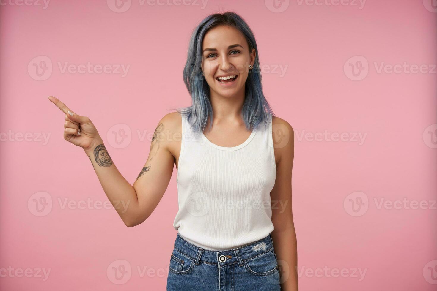 Cheerful young lovely tattooed woman with short blue hair pointing aside with forefinger and smiling widely while standing over pink background in casual wear photo