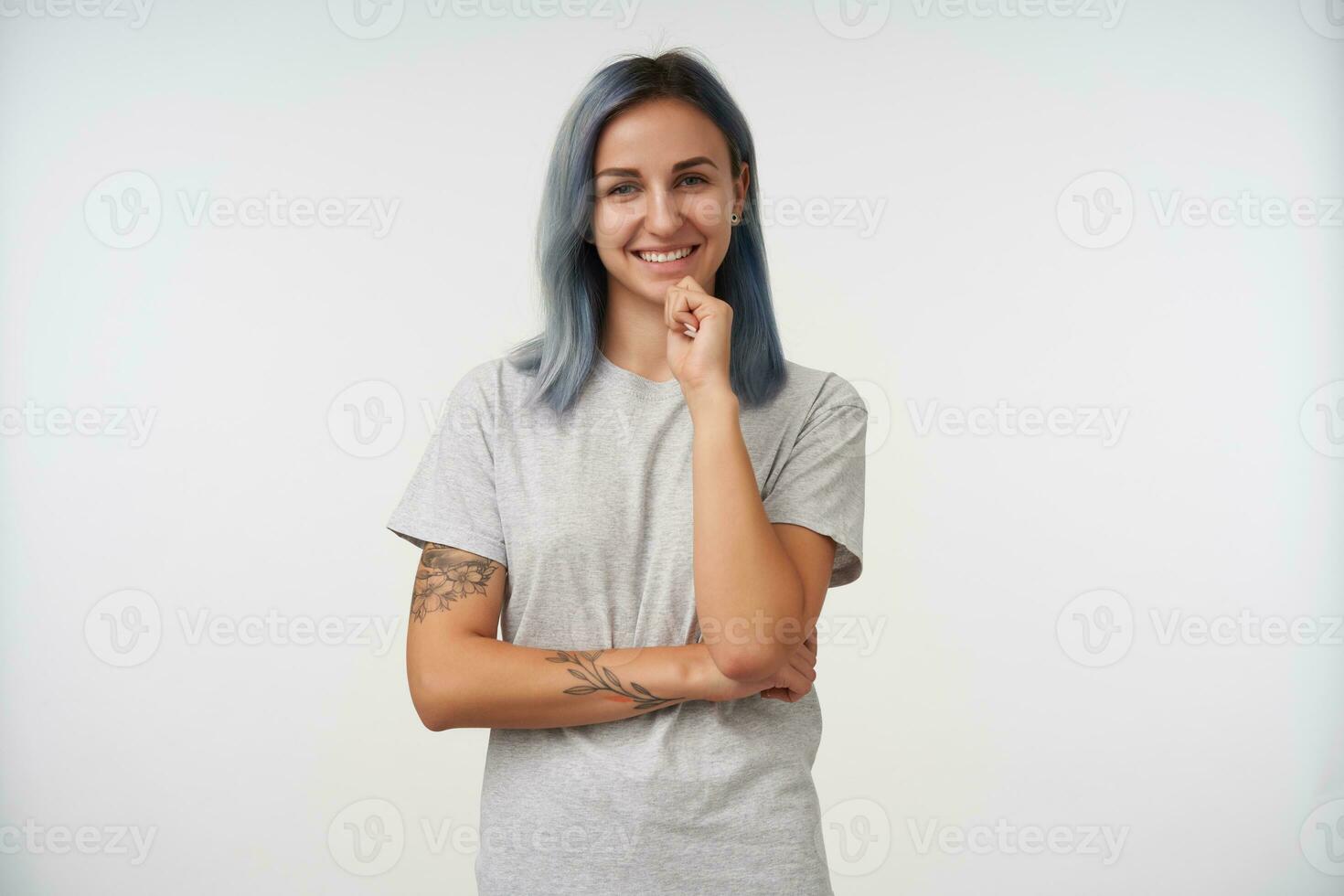 Indoor photo of cheerful young pretty tattooed female with short blue hair holding her chin with raised hand while looking at camera with charming smile, isolated over white background