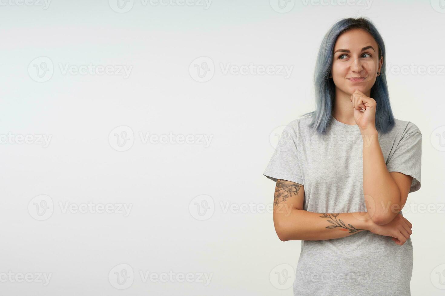 Positive young lovely tattooed woman with short haircut keeping raised hand on her chin while looking thoughtfully aside, standing over white background photo