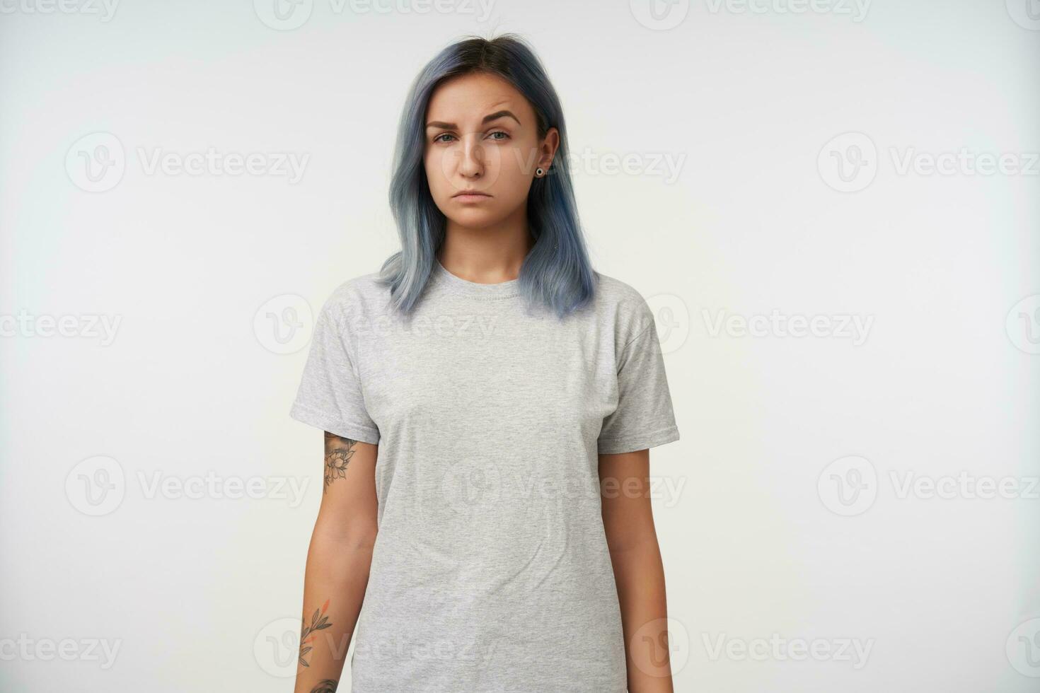 Indoor photo of young attractive tattooed lady squinting her eyes while looking suspiciously at camera and keeping her hands along body while posing over white background