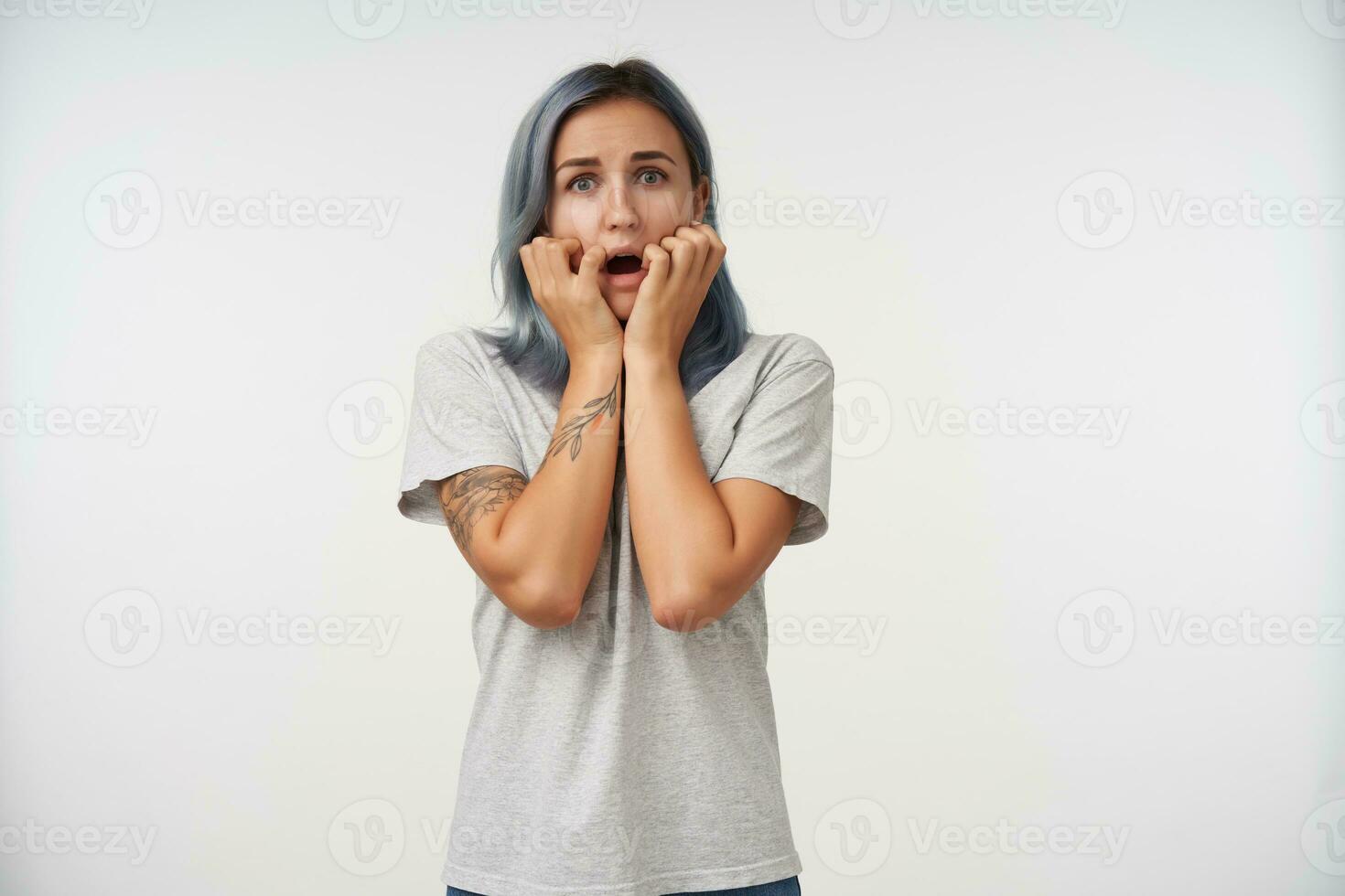 Frightened young attractive woman with short blue hair holding her face with raised hands while looking scaredly at camera, standing over white background photo