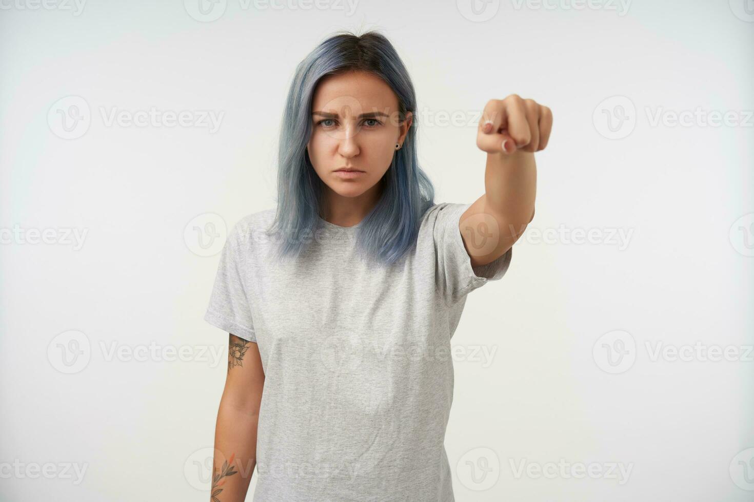 Severe young blue-eyed tattooed female with natural makeup frowning her eyebrows while pointing at camera with forefinger, isolated over white background photo