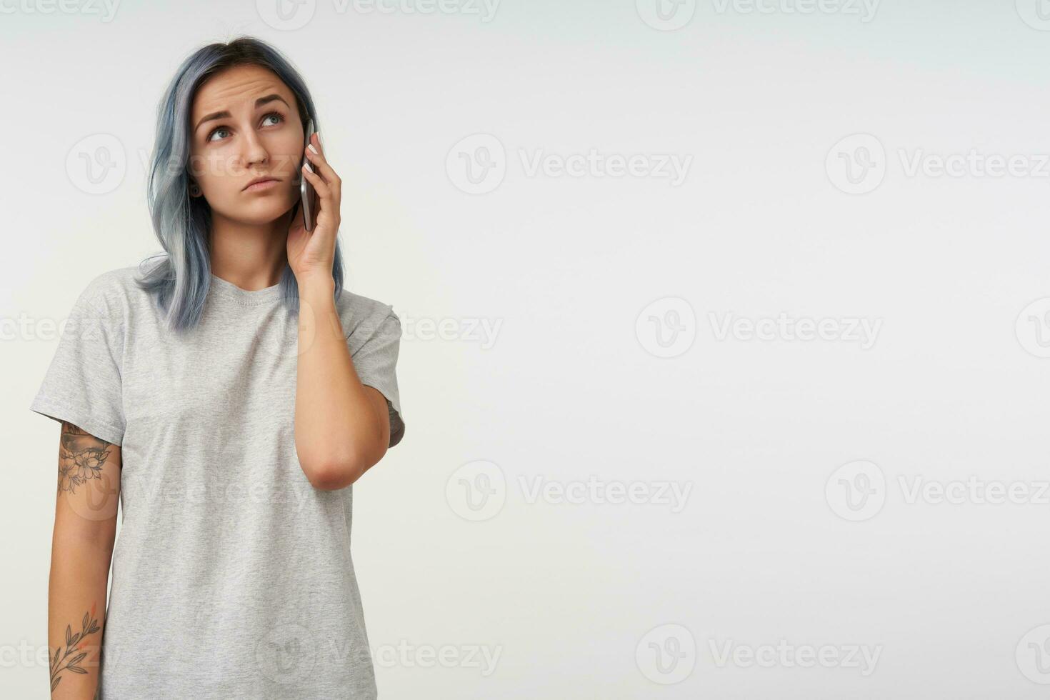 Puzzled young pretty tattooed lady with short blue hair looking pensively upwards while having phone talk, wearing grey basic t-shirt while posing over white background photo