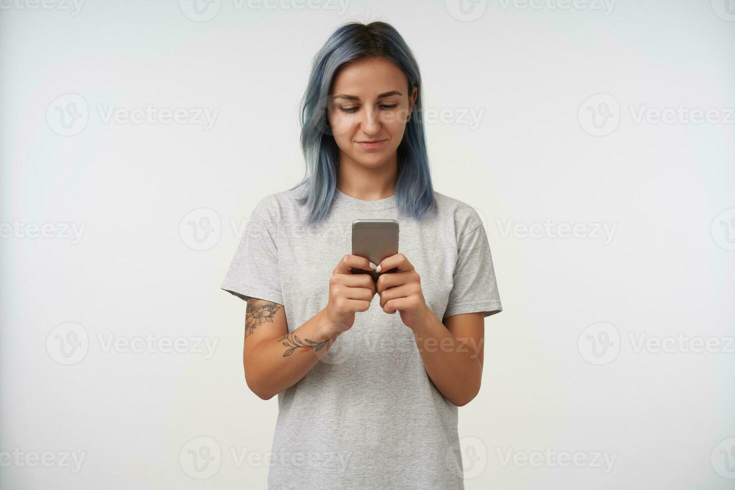 Positive young pretty blue-haired female with tattoos keeping mobile phone in raised hands while checking her messages, isolated over white background photo