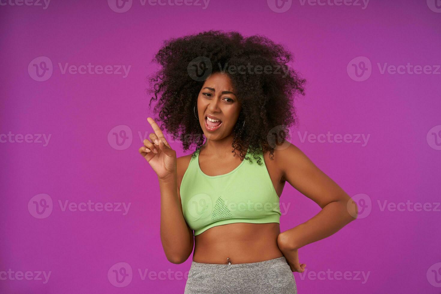 Portrait of young curly woman with dark skin in light green top standing over purple background, looking down wide mouth opened and raising forefinger up photo