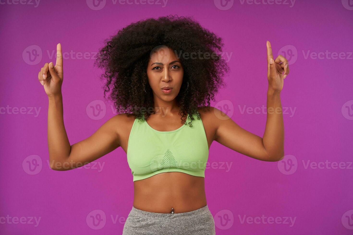 Sporty young dark skinned woman with curly hair lady wearing light green top, showing upwards with forefingers and looking at camera with squint, isolated over purple background photo