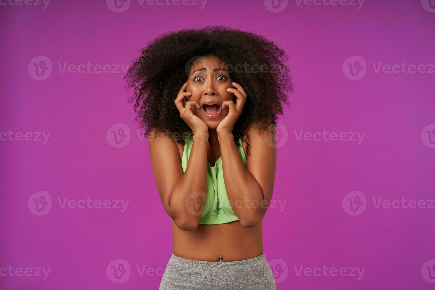 Pop-eyed young curly dark skinned woman standing over purple background with palms on her face, looking scarely to camera with wide mouth opened, wearing casual sporty clothes photo