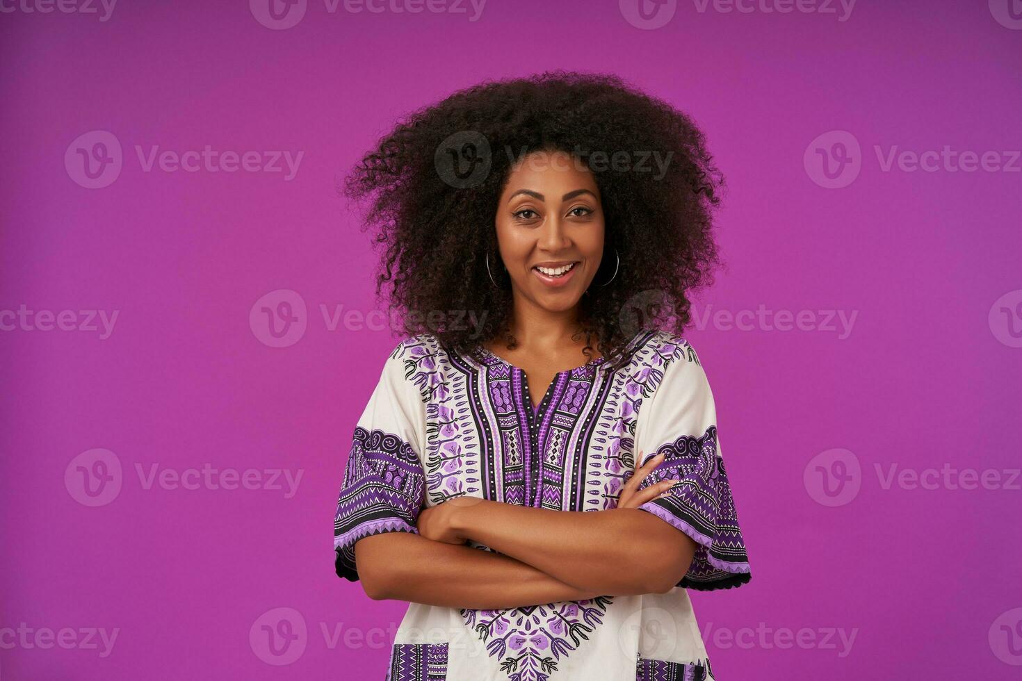 Indoor shot of positive young dark skinned female with curly hair wearing white patterned shirt, posing over purple background with folded hands, looking at camera and smiling cheerfully photo