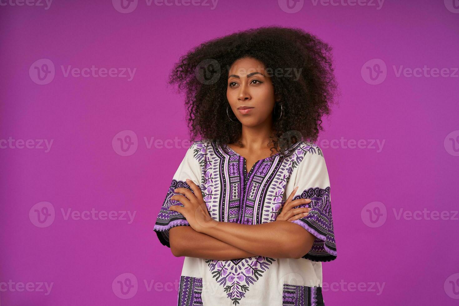 Studio photo on pretty young curly female with dark skin holding folded hands on her chest, looking aside with serious face, isolated over purple background in casual wear