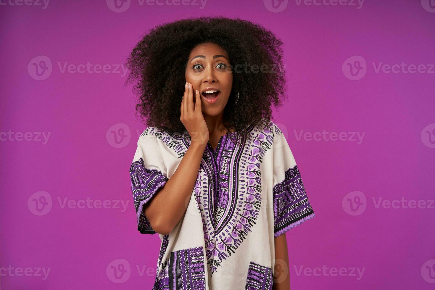 Inoor photo of amazed young dark skinned woman with curly hair keeping raised palm on her face and looking at camera with suprised face, isolated over purple background