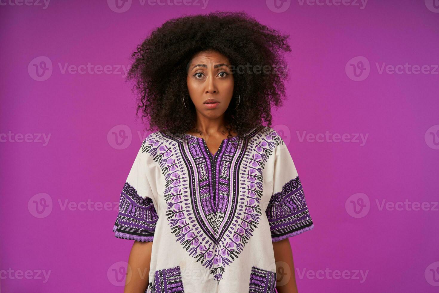 Portrait of upset curly dark skinned female in casual clothes standing over purple background wth hands down, contracting forehead and looking to camera with sad face photo