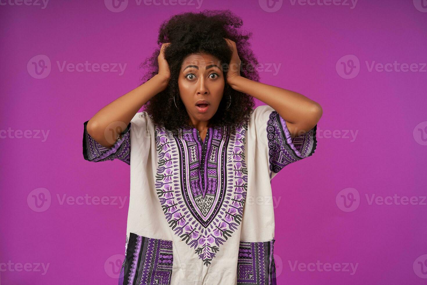 Scared young dark skinned woman with curly hair wearing white patterned shirt, holding her head with raised hands and looking at camera with wide eyes and mouth opened, isolated over purple background photo