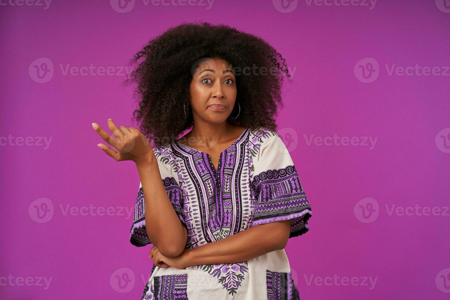 Portrait of young curly lady casual with dark skin in white patterned shirt rasing palm confusedly, looking at camera with raised eyebrows while standing over purple background photo
