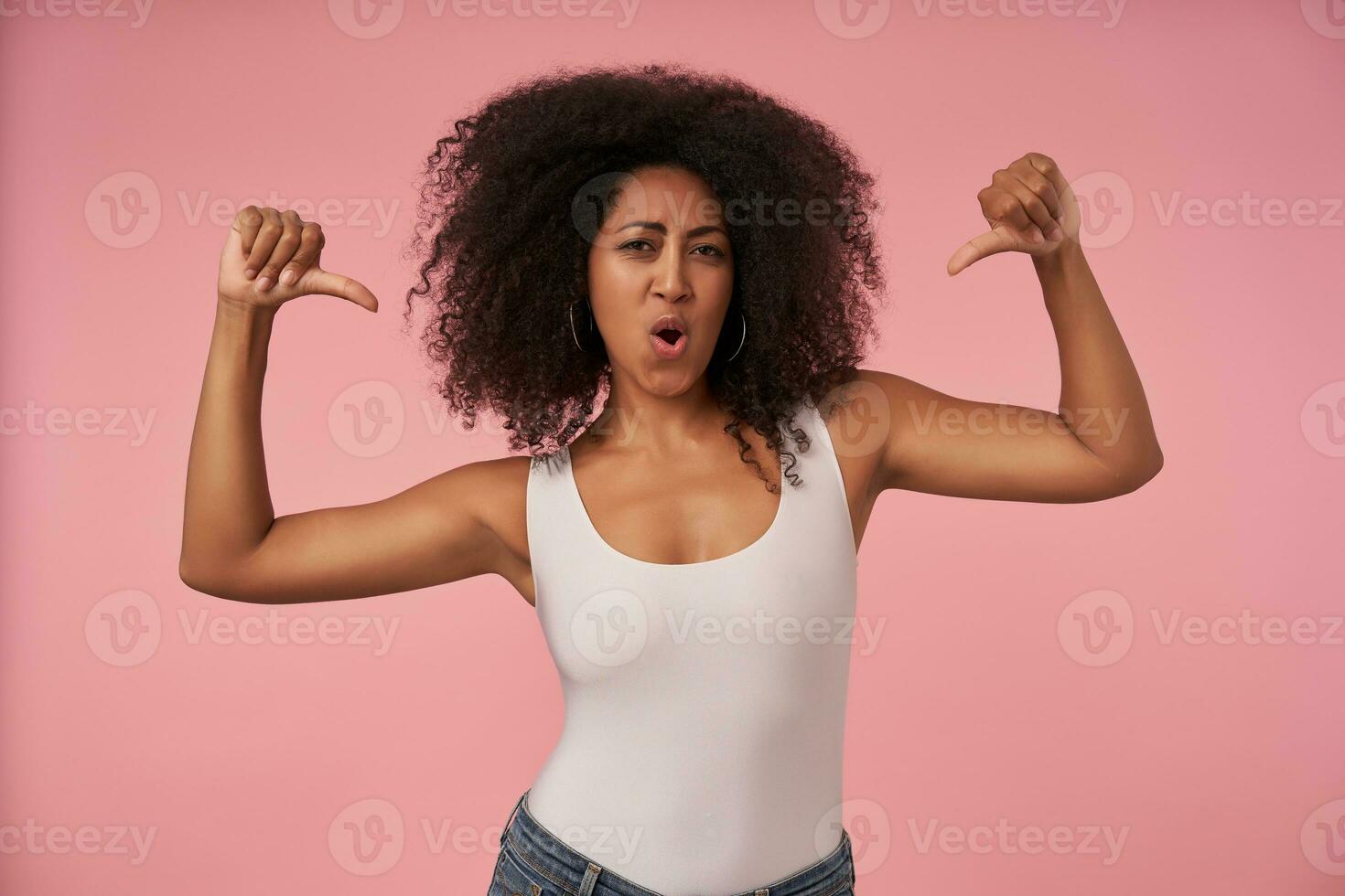 Studio shot of young curly dark skinned lady in casual clothes looking at camera self-confidently, posing over pink background with raised thumbs showing down photo