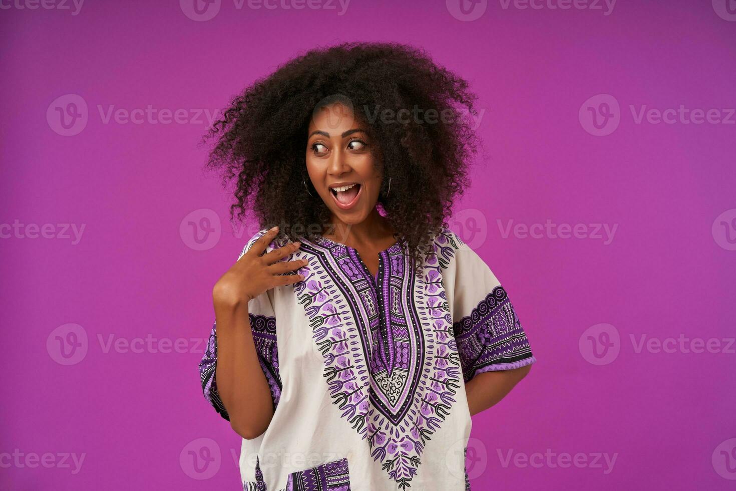 Joyful young dark skinned woman with casual hairstyle keeping palm on her shoulder and looking aside with wide joyful smile, posing over purple background photo