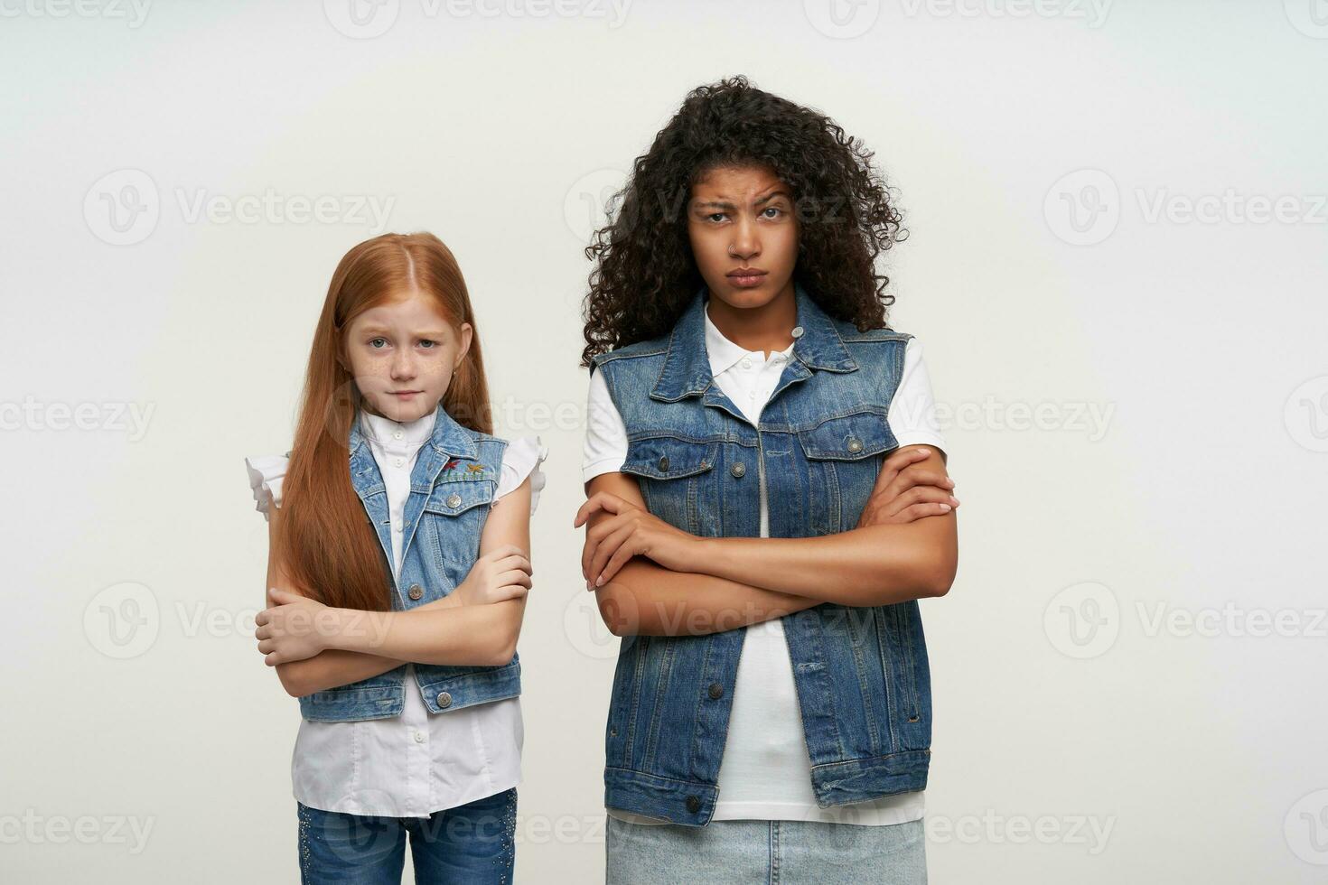 Couple of little redhead girl with long hair and young curly brunette female with dark skin raising their eyebrows and looking to camera with displeased face, standing over white background photo