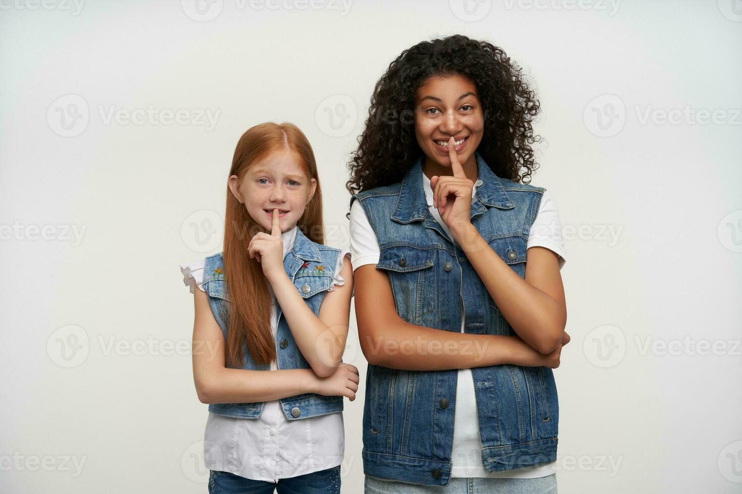 Cheerful couple of young dark skinned curly brunette lady and redhead long haired pretty girl keeping forefingers on their lips, asking to keep secret and smiling cunningly photo