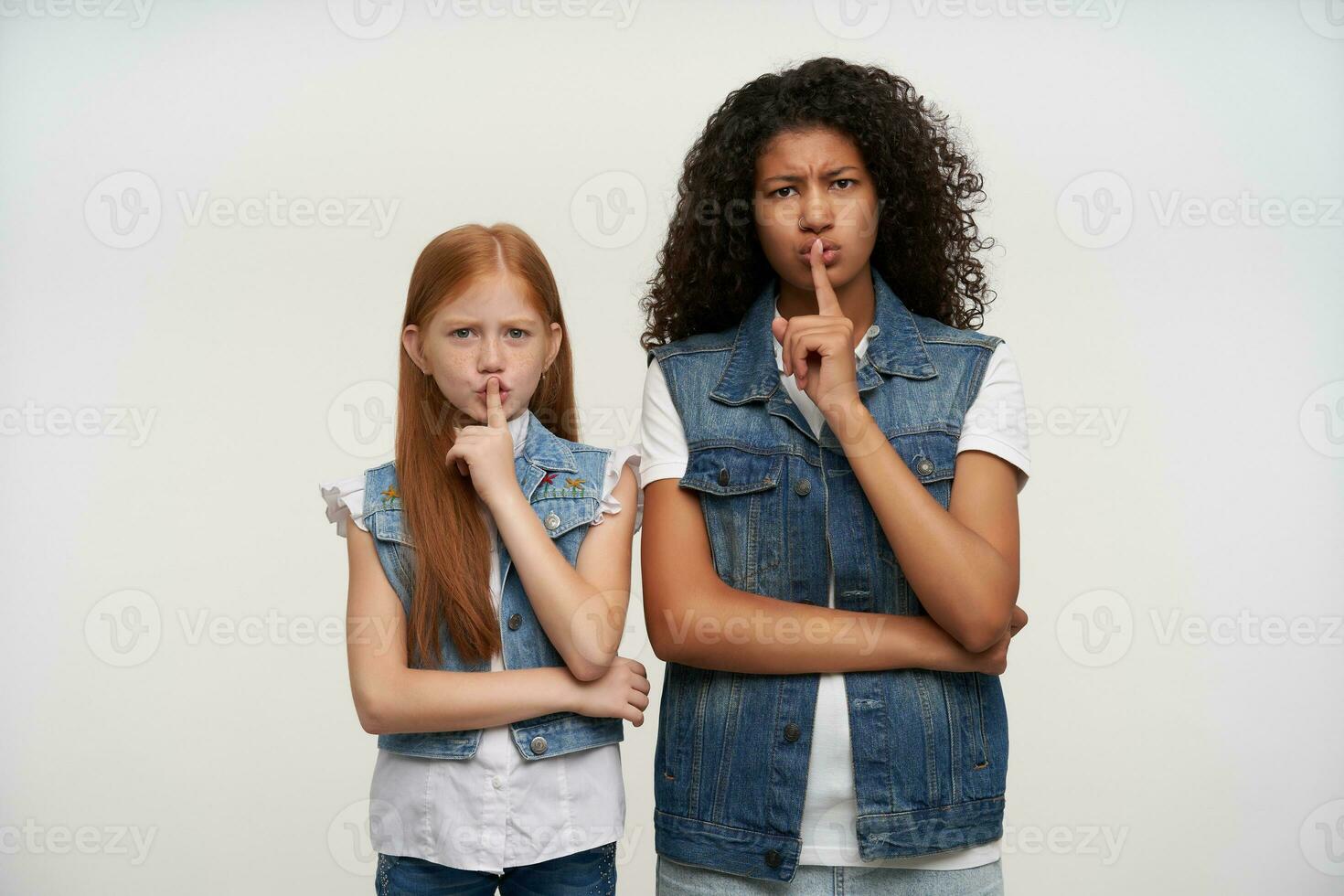 Serious looking young long haired ladies frowning their eyebrows and raising forefingers in hush gesture, asking to keep silence, wearing casual clothes while posing over white background photo