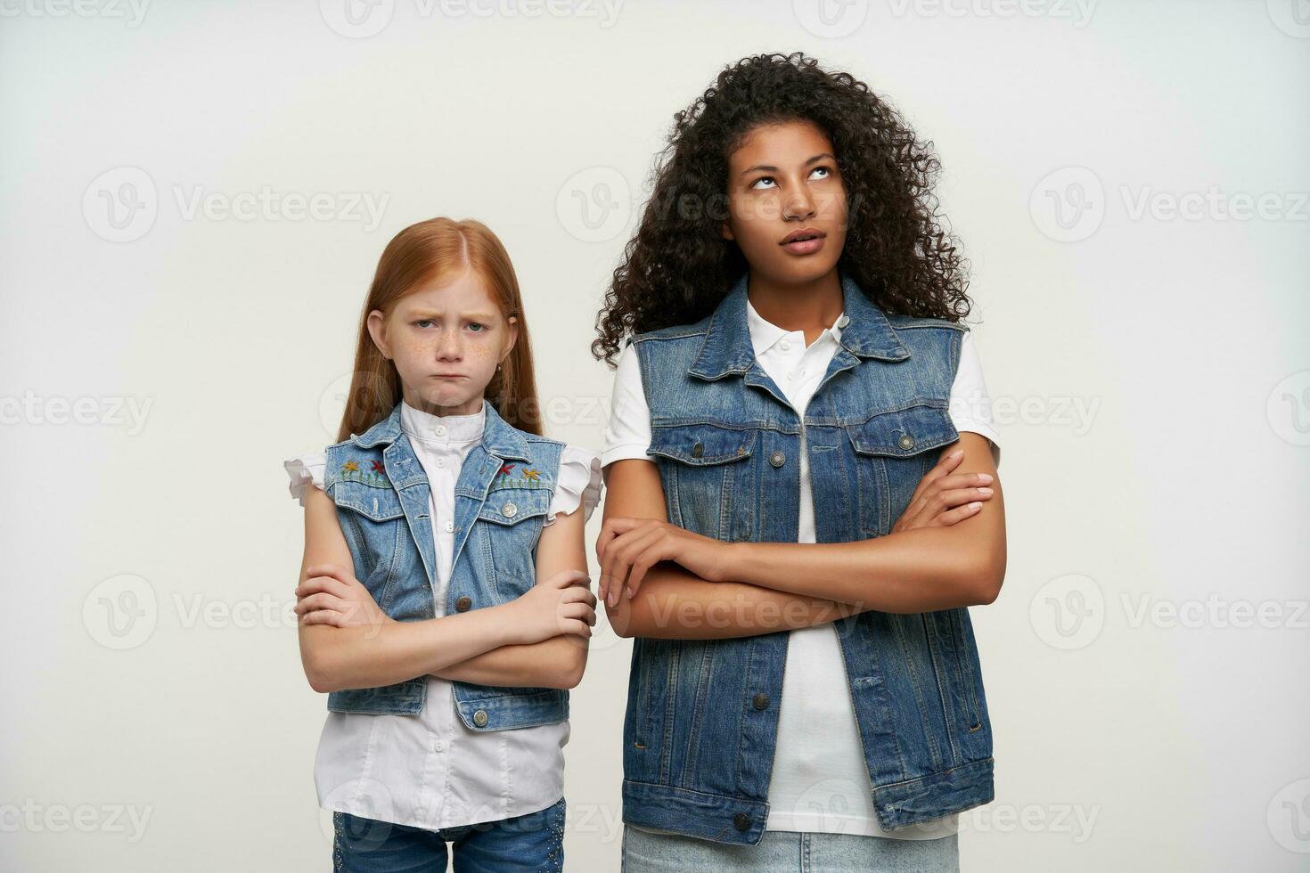 Couple of offended young pretty girls posing over white background, keeping their hands folded and standing with displeased faces, having quarrel and do not want to talk to each other photo