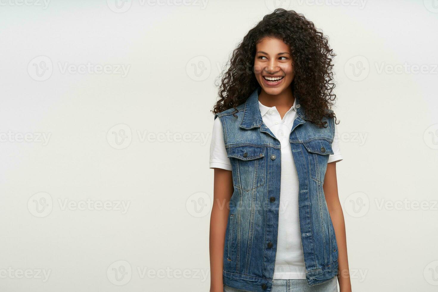 Cheerful pretty young brunette curly woman with dark skin standing over white background with hands down, looking aside happily with charming smile, being in high spirit photo