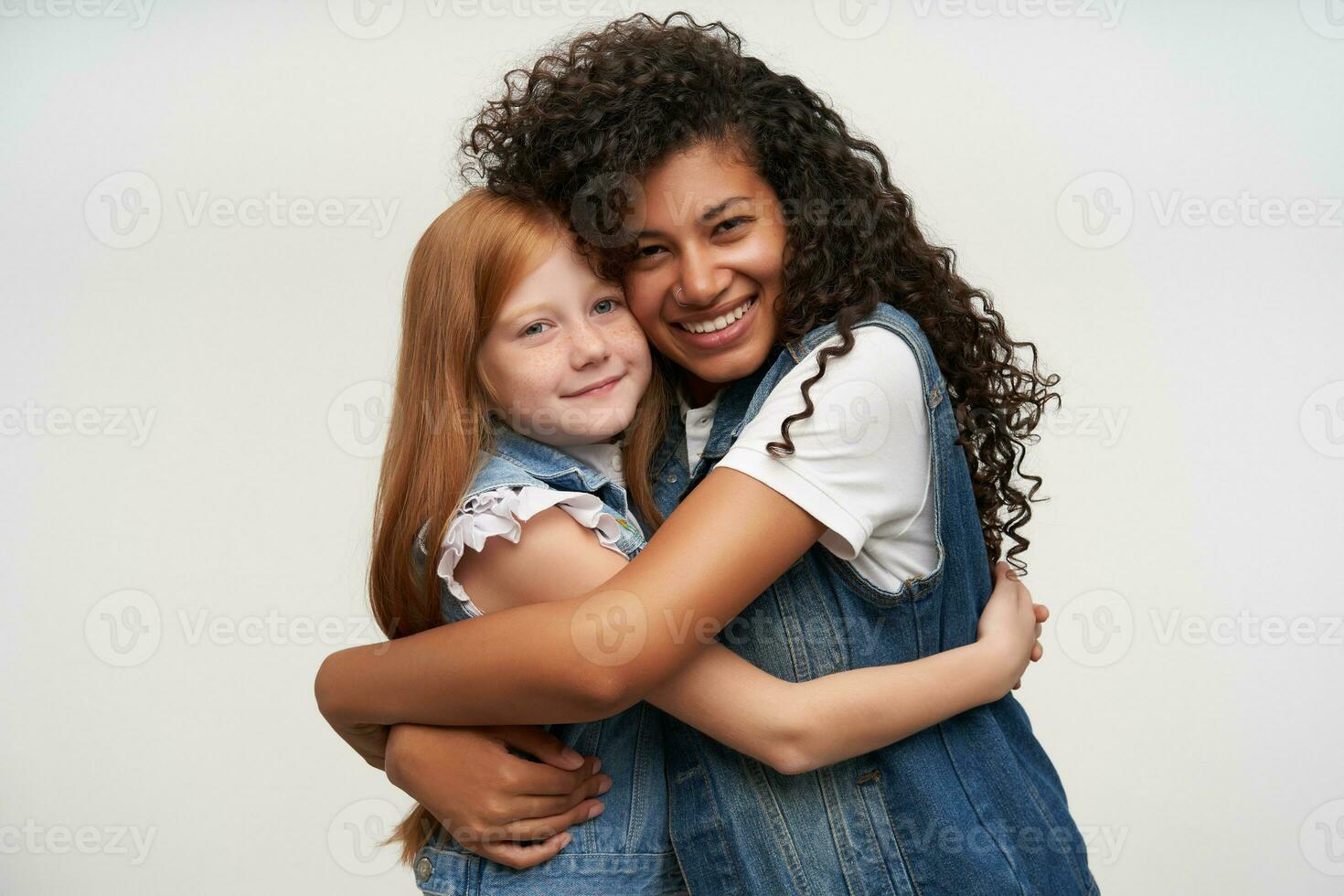 Portrait of attractive happy young dark skinned woman with long curly hair hugging pretty red haired little girl and smiling widely while posing over white background photo