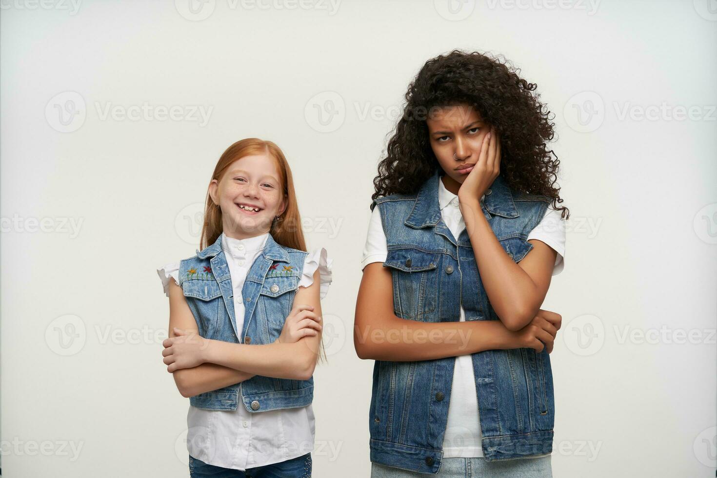 Gloomy young curly brunette female with dark skin looking sadly at camera and leaning her head on raised hand, posing over white background with cute cheerful redhead little girl photo