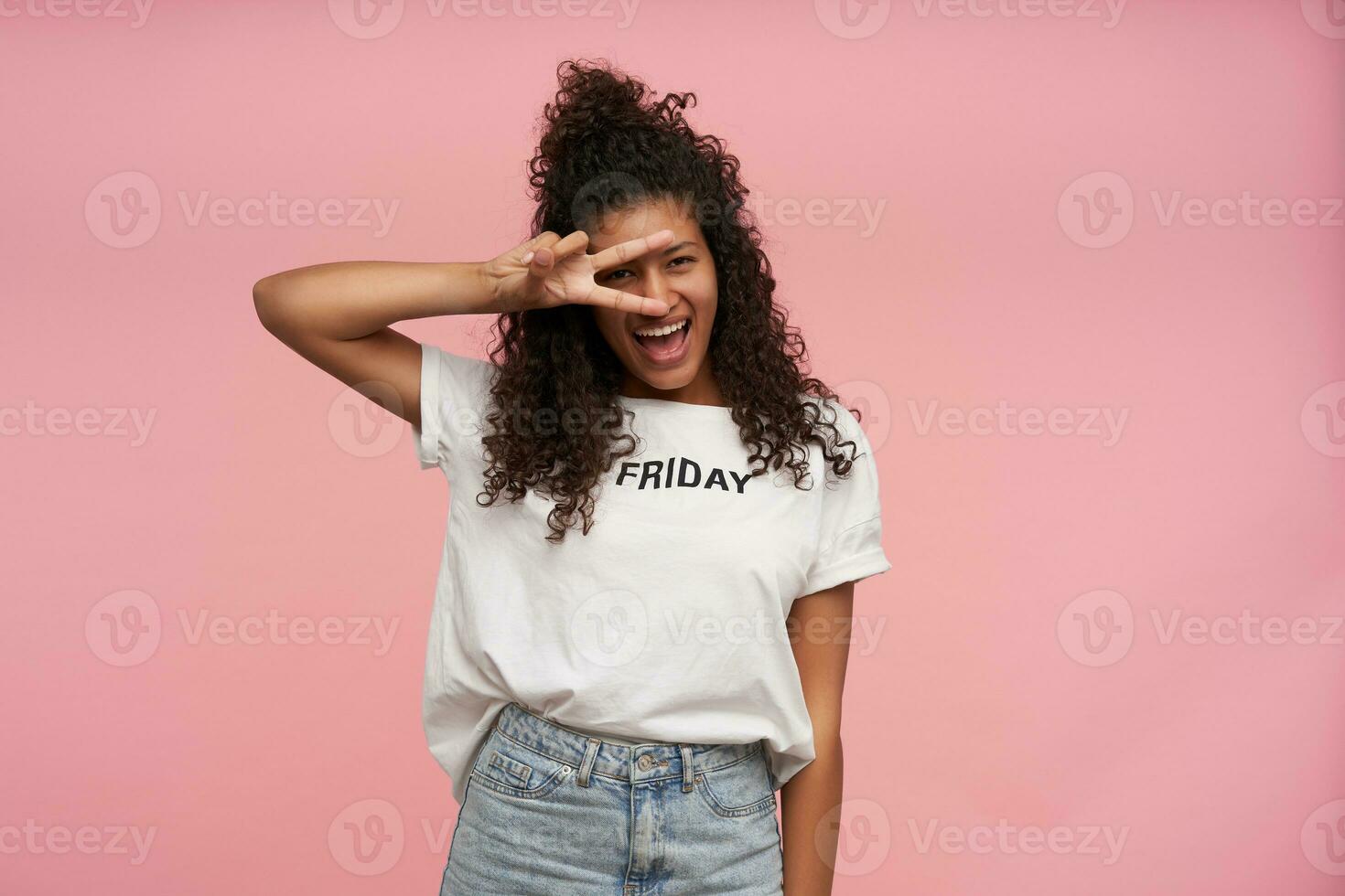 Indoor shot of happy young brunette dark skinned female with curly long hair dressed in white t-shirt and blue jeans posing over pink background, raising peace gesture to her face and smiling joyfully photo