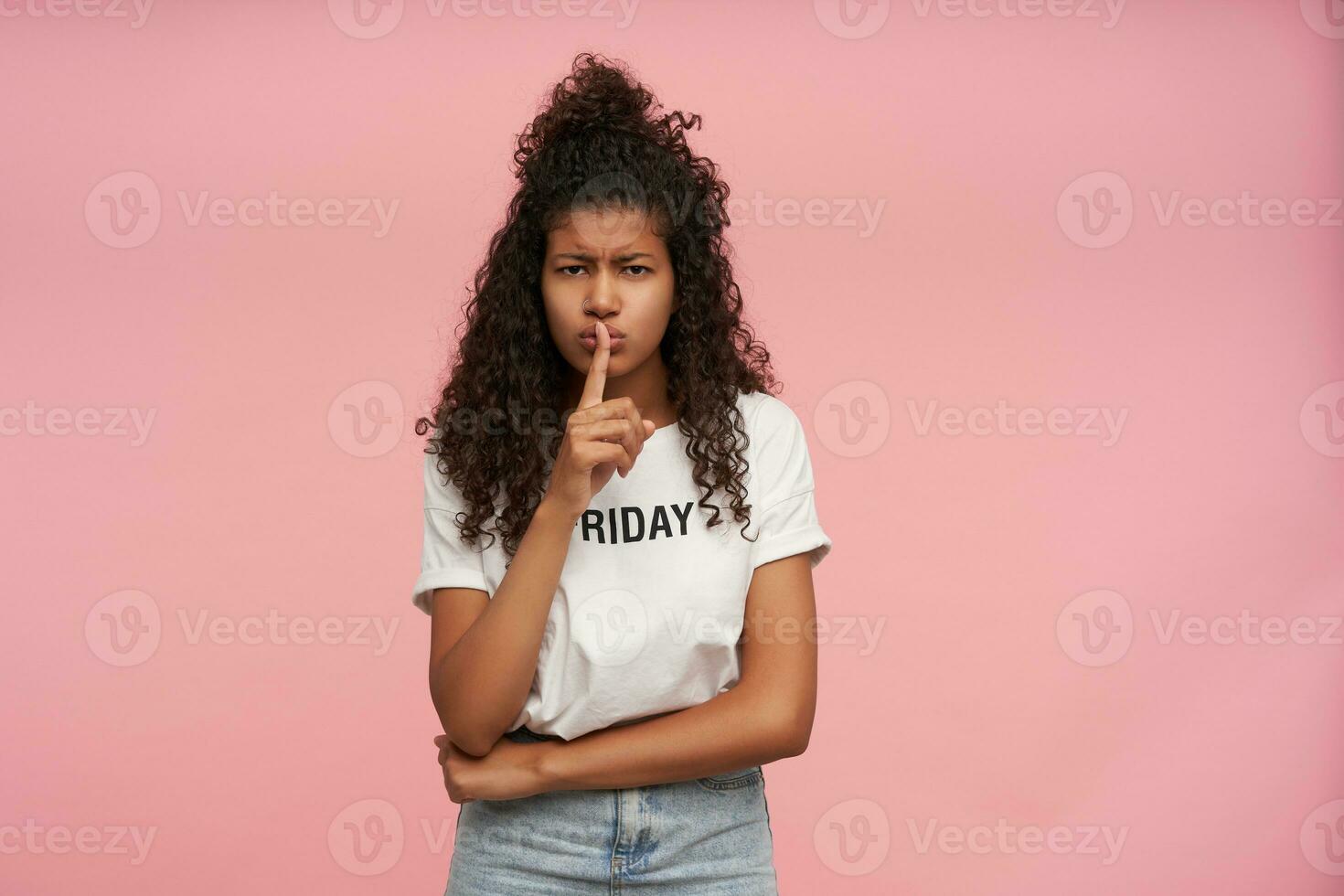 Portrait of serious looking young curly long haired brunette lady with dark skin frowning eyebrows and holding forefinger on her lips, asking to keep silence, isolated over pink background photo