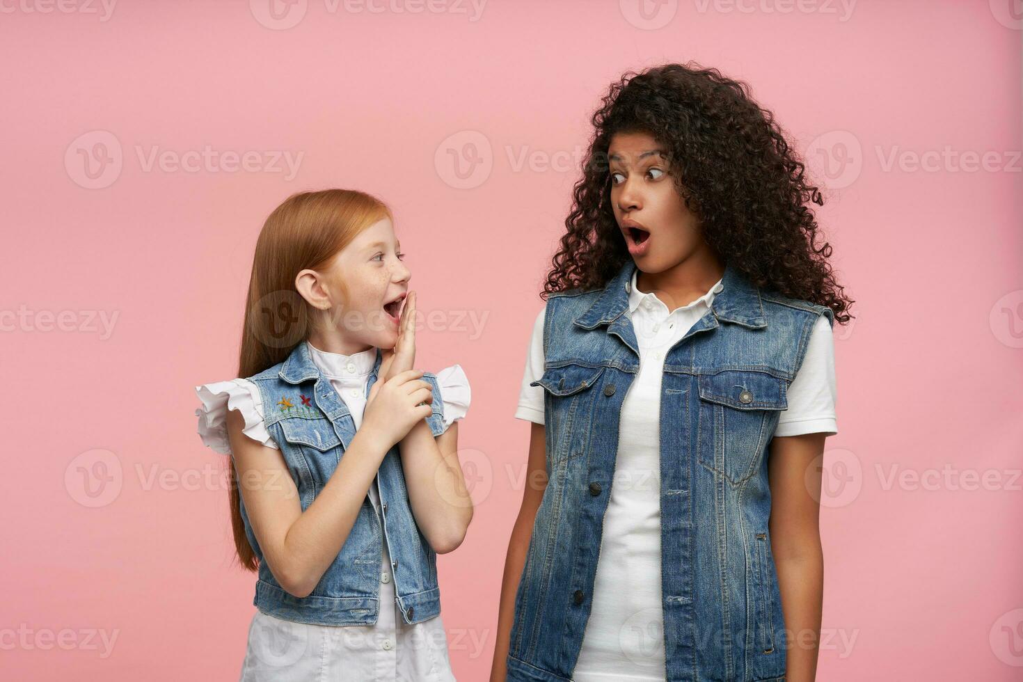 Indoor photo with young couple of shocked pretty girls looking amazedly on each other with wide mouth opened and rounding surprisedly eyes, isolated over pink background