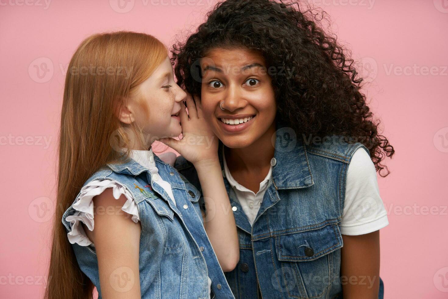 Close-up of pretty young dark skinned curly brunette woman with nose piercing looking wonderingly at camera and raising eyebrows surprisedly while listening to secret stories over pink background photo