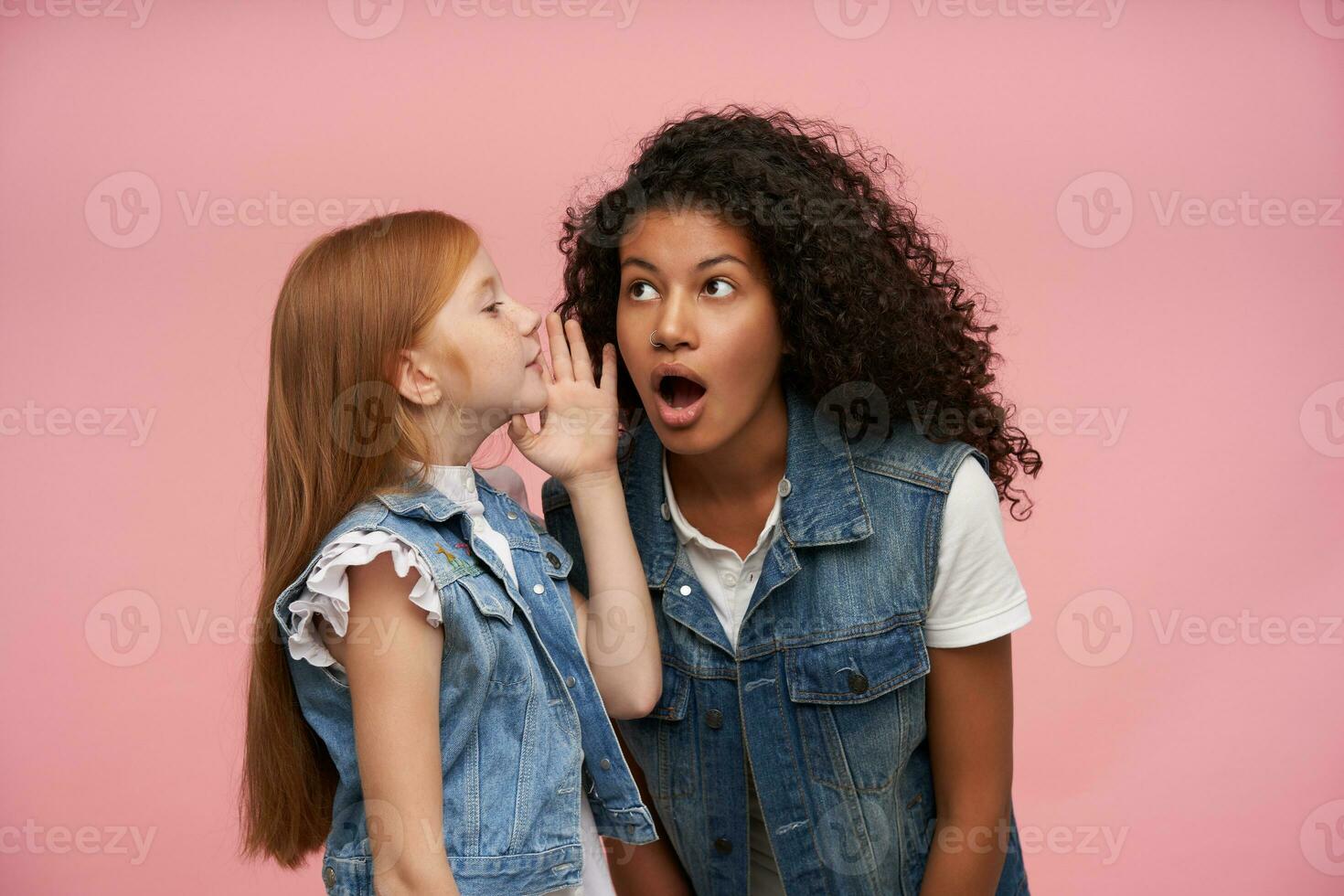 Portrait of amazed young dark skinned brunette female with long curly hair listening to excited news and keeping her mouth wide opened, posing against pink background photo