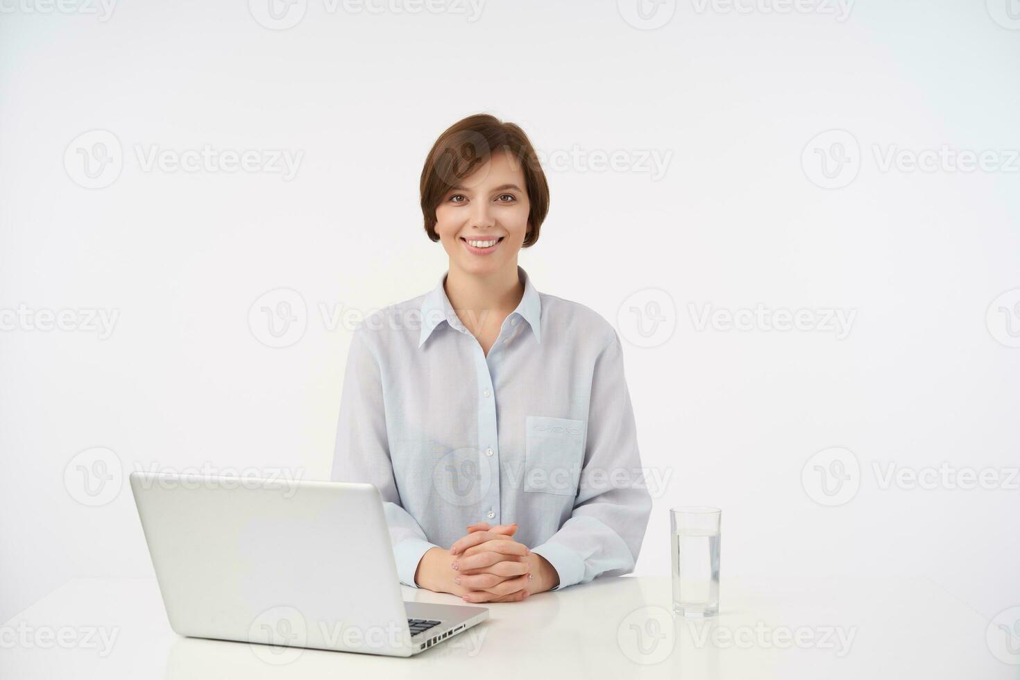 positivo joven bonito corto peludo morena hembra sentado a mesa y plegable manos en encimera, mirando alegremente a cámara con encantador sonrisa, aislado terminado blanco antecedentes foto