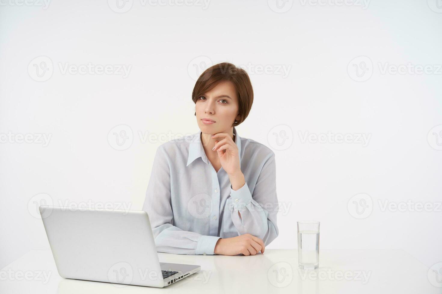 Serious young brown-eyed short haired female with natural makeup keeping raised hand under her chin and looking pensively at camera with folded lips, sitting at table over white background photo