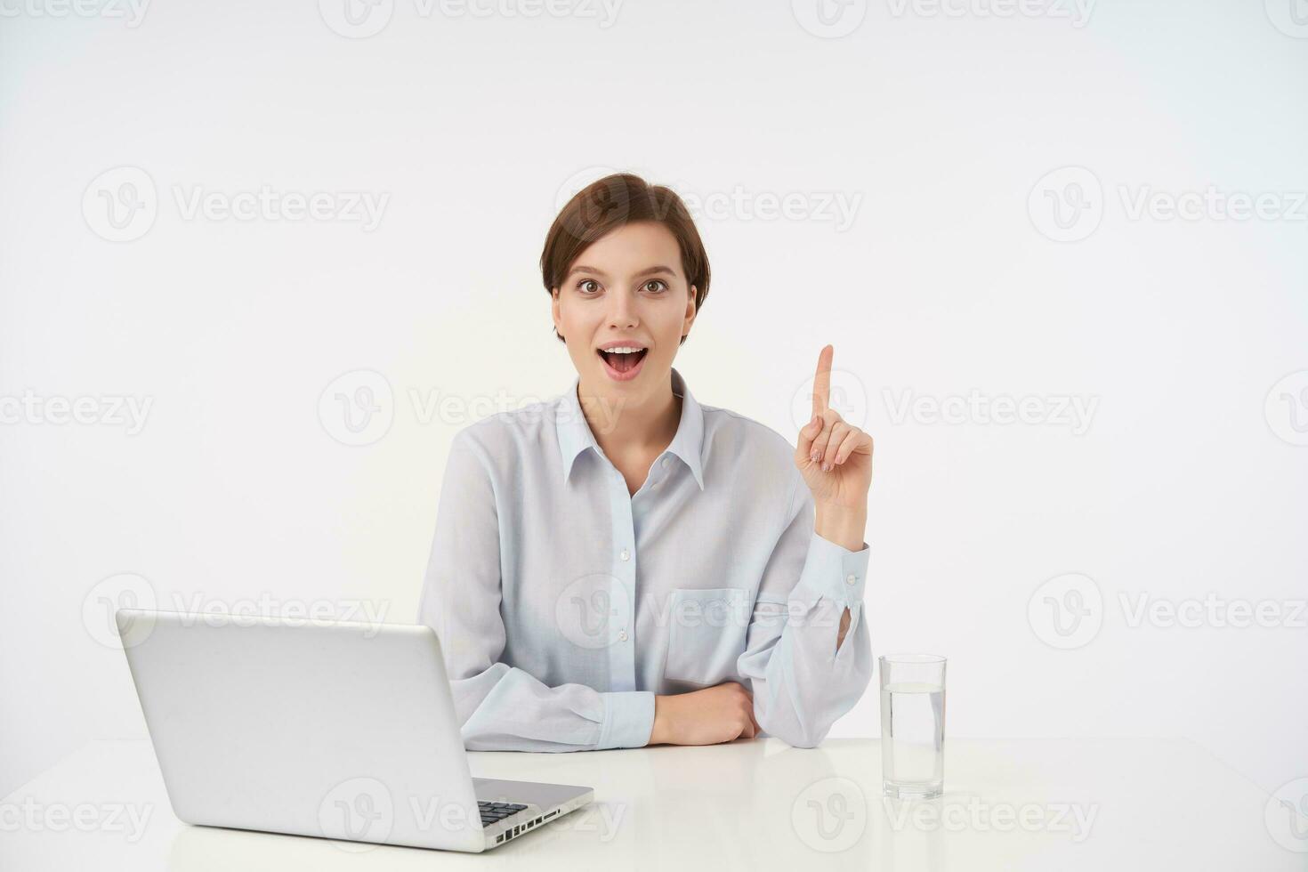 Joyful young pretty short haired brunette female with casual hairstyle sitting over white background and showing upwards with raised index finger, looking cheerfully at camera with wide mouth opened photo