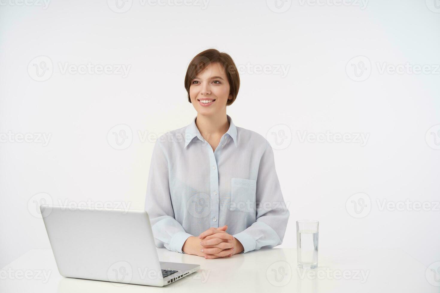 Cheerful attractive young brown haired woman employee sitting over white background with modern laptop and being ready for meeting, looking aside with positive smile photo