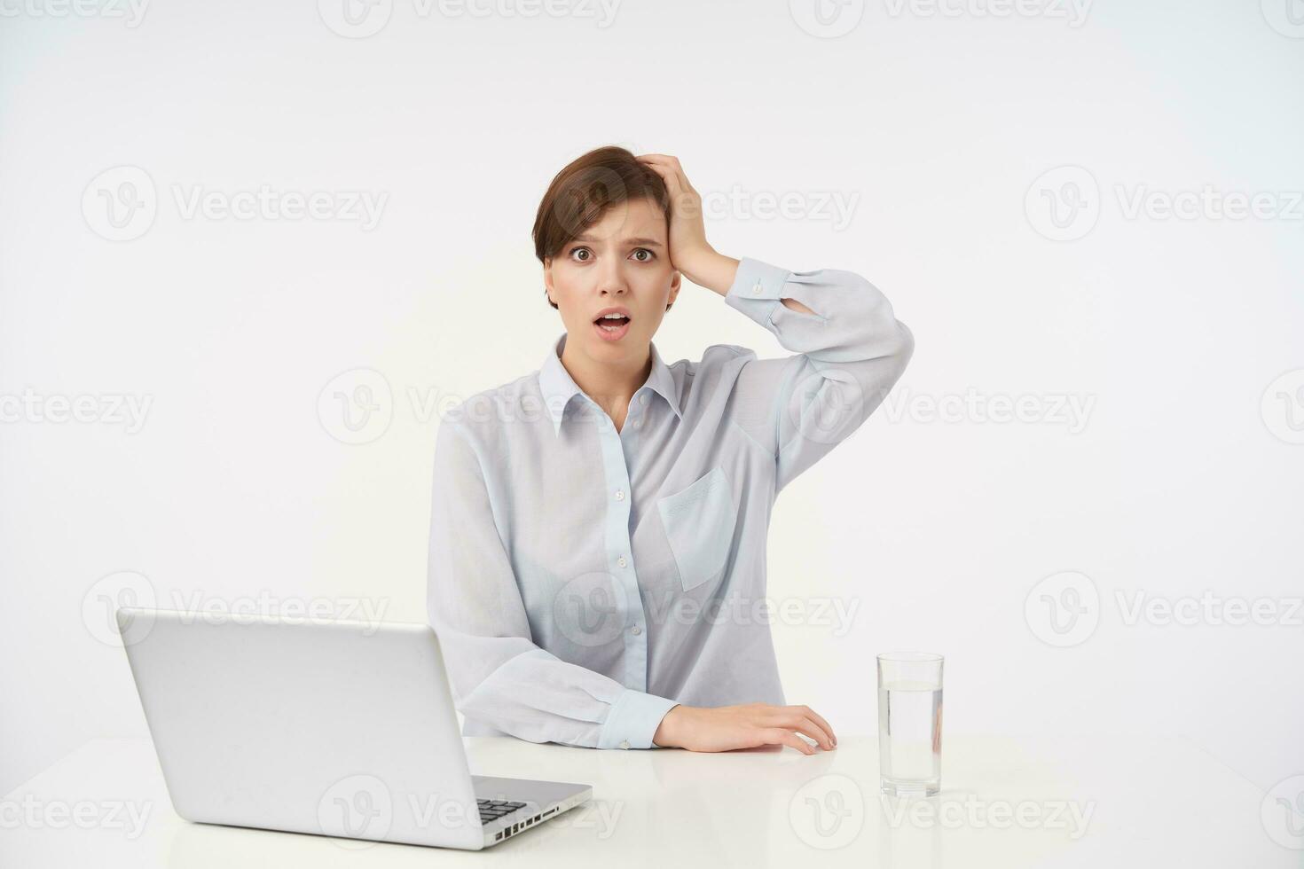 Puzzled young attractive short haired brunette lady keeping palm on her head and looking confusedly at camera, dressed in formal clothes while sitting over white background photo