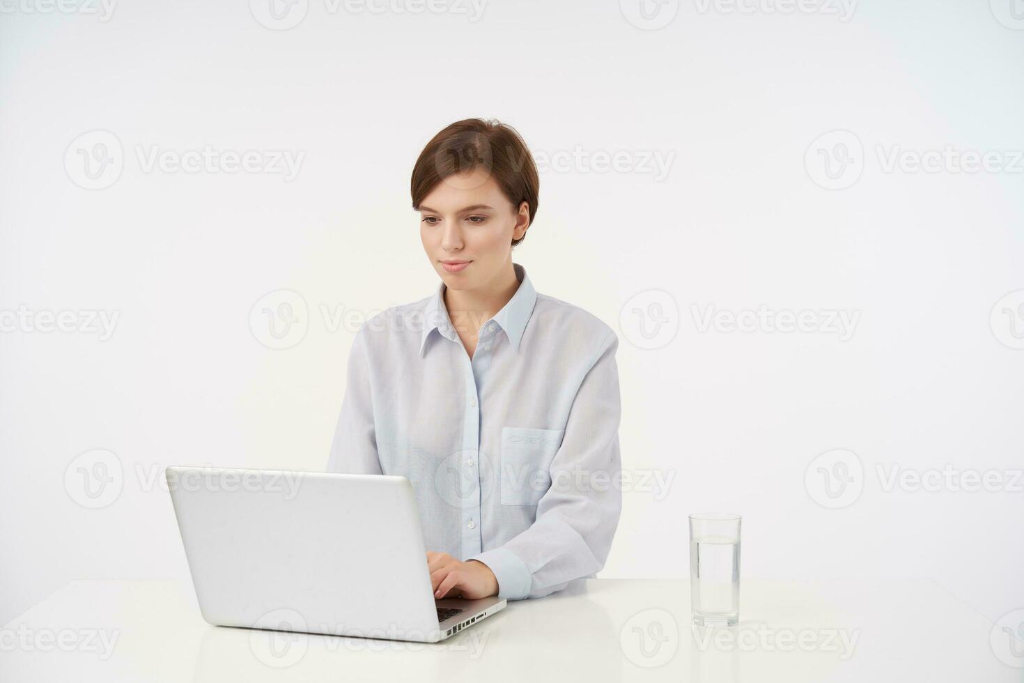 Indoor photo of charming young short haired brunette lady with casual hairstyle holding her hand on keyboard and typing text on laptop, isolated over white background