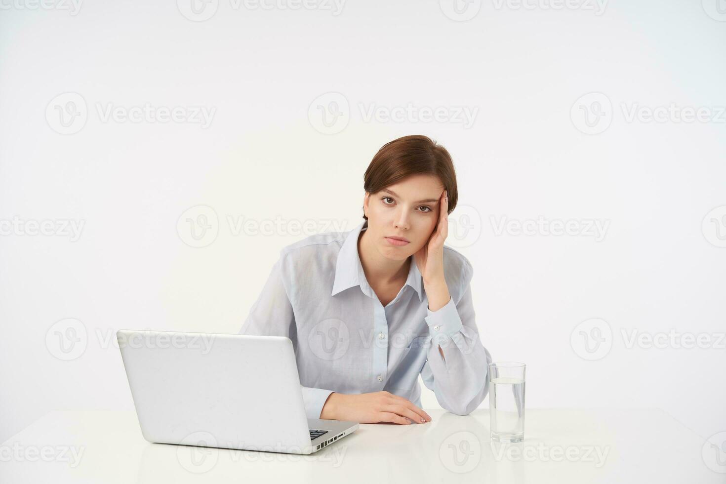 Exhausted young pretty short haired brunette lady with natural makeup leaning her head on raised hand and looking tiredly at camera, isolated over white background photo