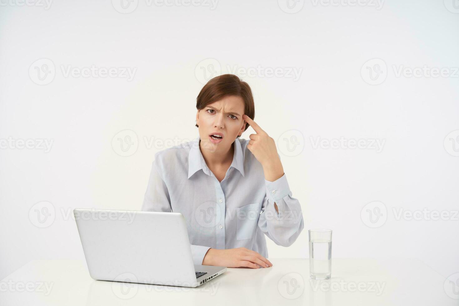 Indoor photo of young dissatisfied brown haired female folding gun with raised hand and keeping it on her temple, frowning eyebrows with pout while looking at camera