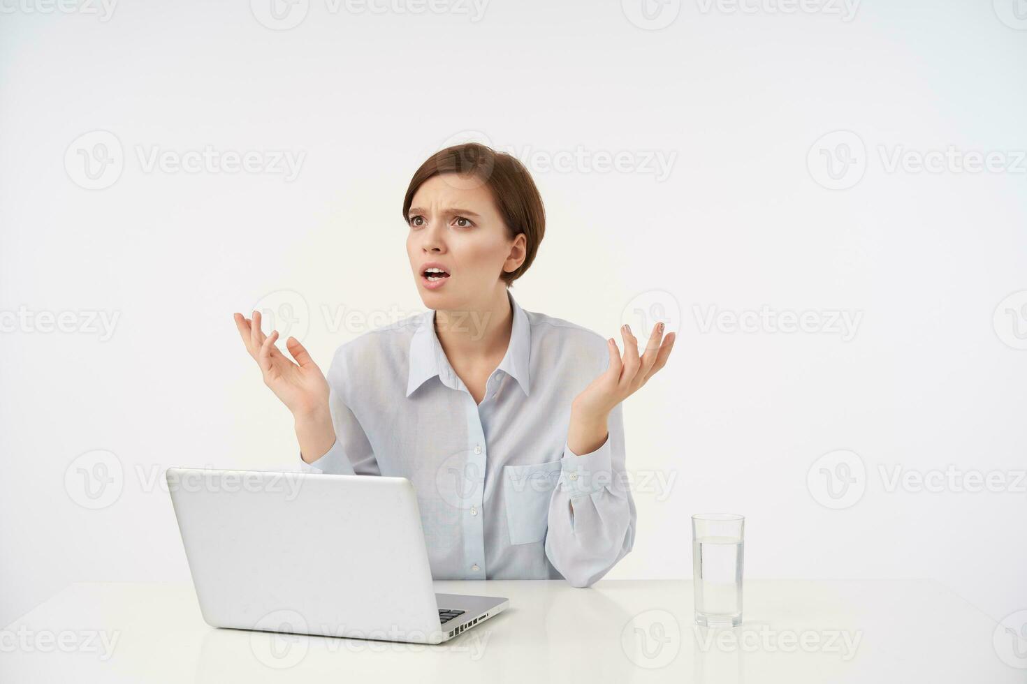 Bewildered young brown haired pretty woman with casual hairstyle frowning her eyebrows and raising confusedly palms while looking aside, sitting at table over white background photo
