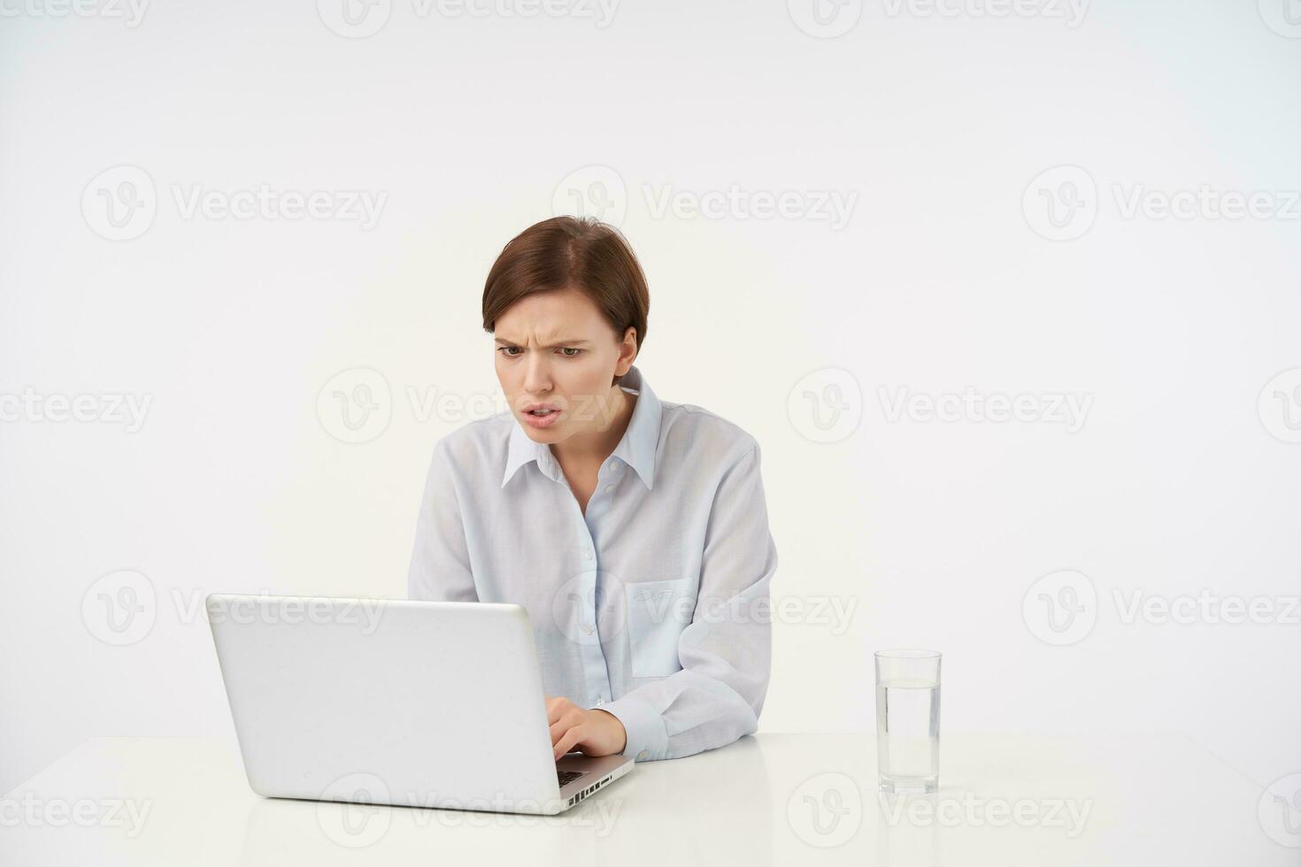 Shot of confused young short haired brunette lady with casual hairstyle looking at screen of her laptop with displeased face, reading unpleasant news, isolated over white background photo