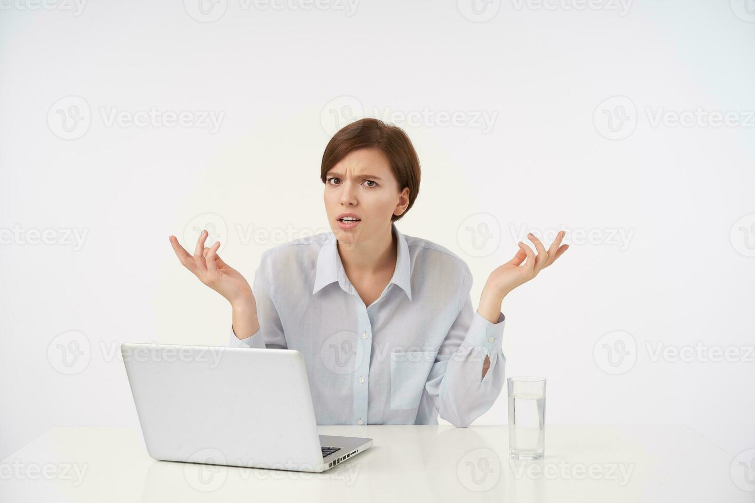 Puzzled young brown-eyed short haired woman with natural makeup looking at camera with displeased face and raising confusedly palms while sitting over white background photo