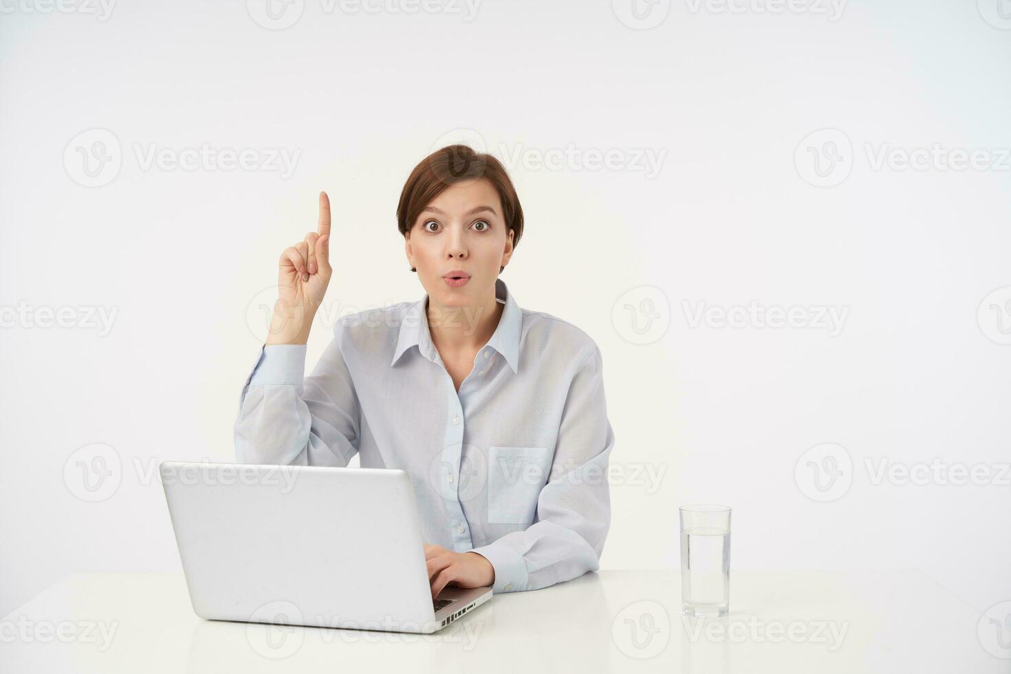 Excited young brown-eyed short haired brunette lady with natural makeup looking at camera surprisedly while posing over white background in formal clothes photo