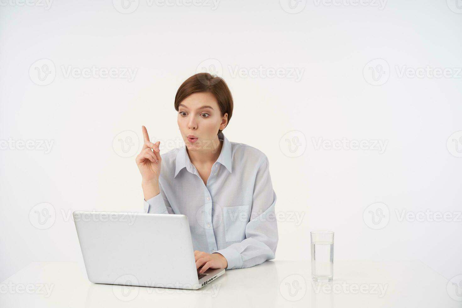 Indoor photo of attractive young short haired brunette female rounding excitedly her mouth and raising forefinger as she is having good idea, posing over white background