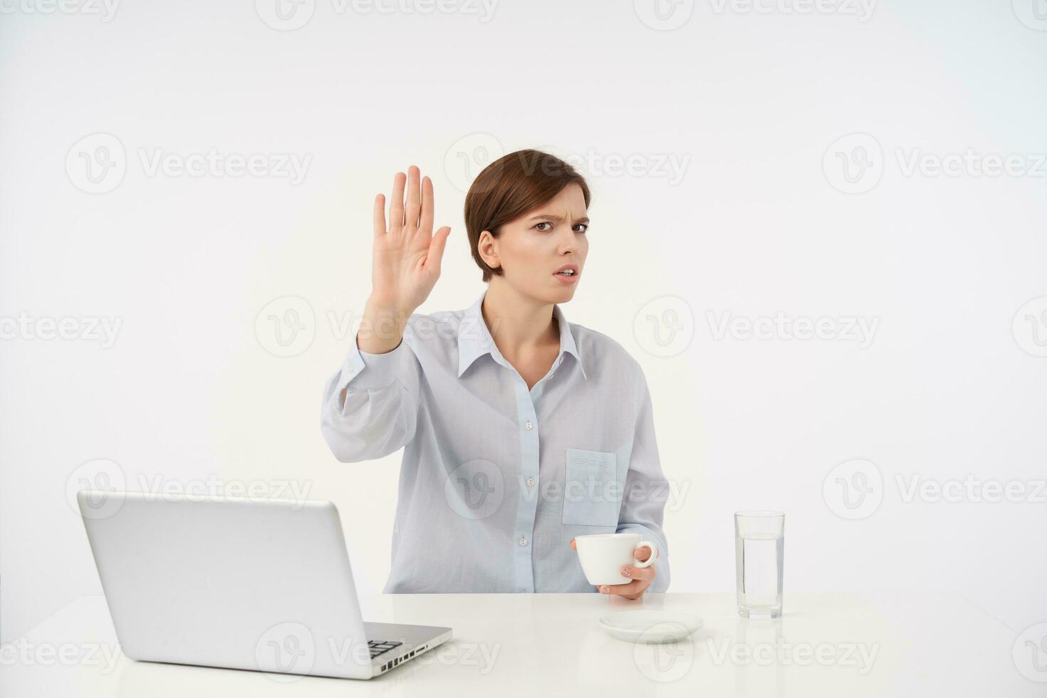 Indoor photo of young brown haired woman with casual hairstyle holding ceramic cup and raising palm in stop gesture, frowning eyebrows with displeased face while sitting over white background