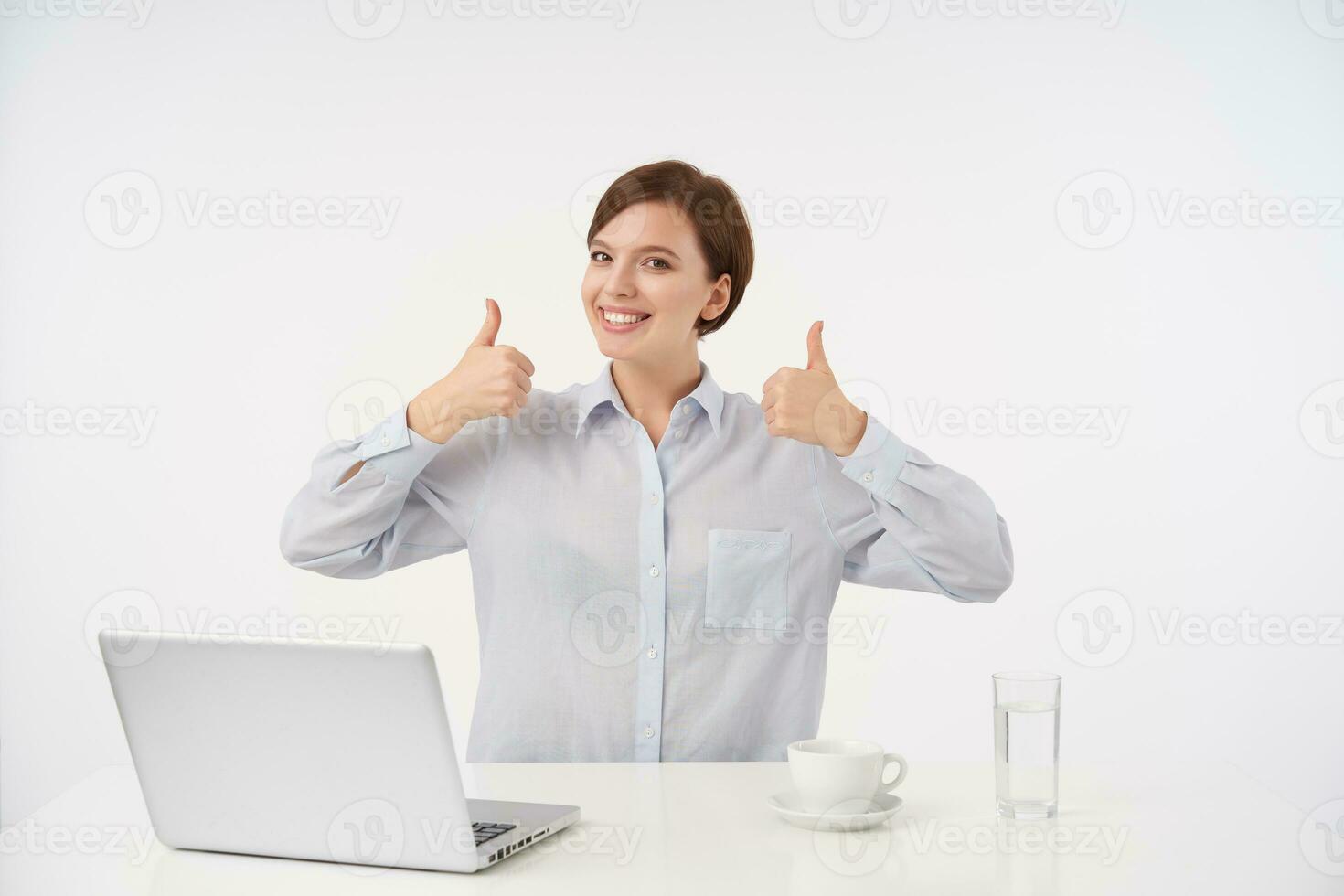 Cheerful young lovely brown haired woman with natural makeup looking happily at camera and showing raised thumbs at camera, wearing formal clothes while posing over white background photo